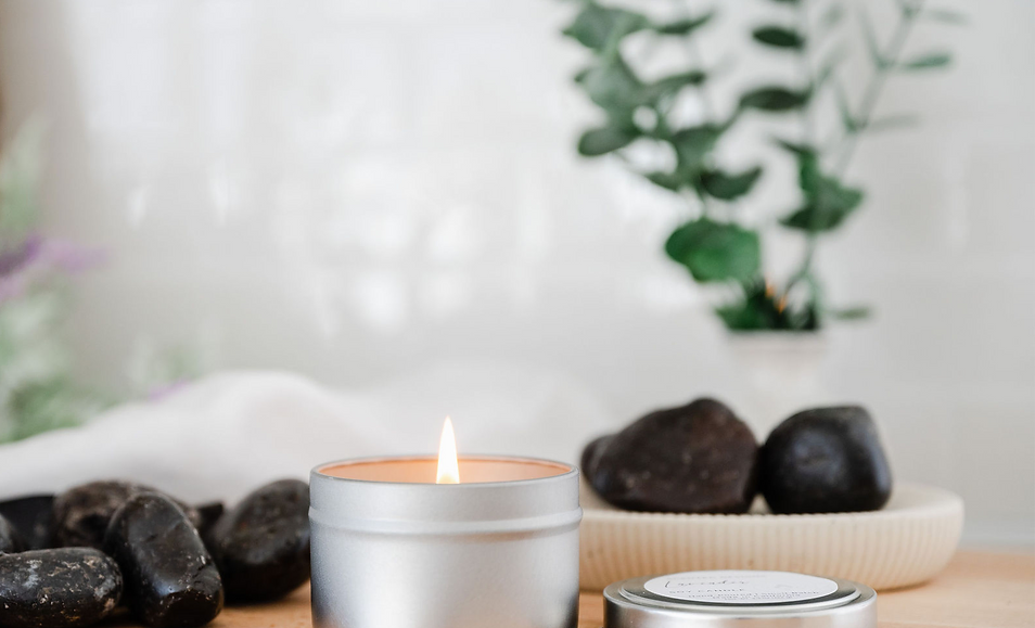 A metal tin candle is burning on a kitchen counter. In the background are spa rocks, lavender, and eucalyptus for a calming atomsphere