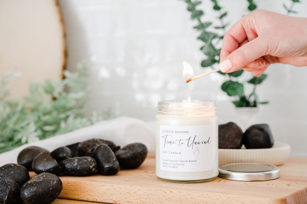A soy candle sits on a kitchen counter. Spa rocks and eucalyptus are behind it. The soy wax candle is being lit by a match. 